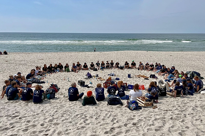 Großer Kreis aus etwa 60 im Sand sitzenden Personen 